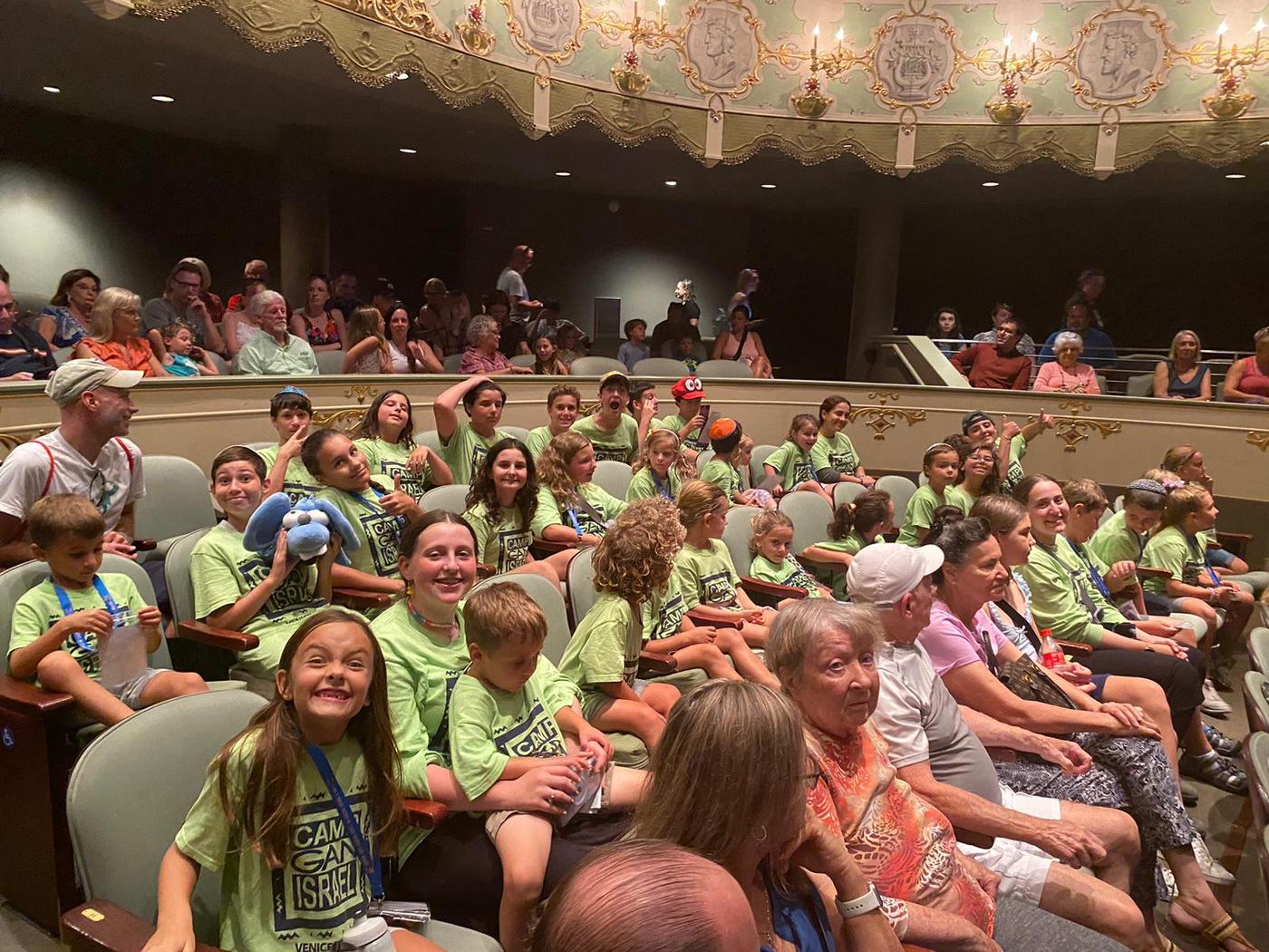 Camp Gan Israel campers and counselors in green camp shirts filling rows of theater seats on a trip day