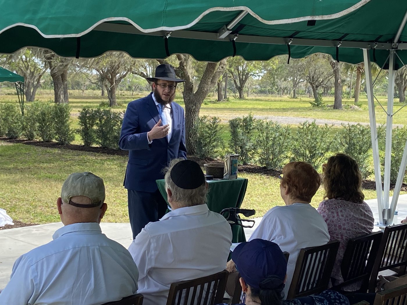 A rabbi leading a graveside service under a canopy at Jerusalem Gardens, family seated