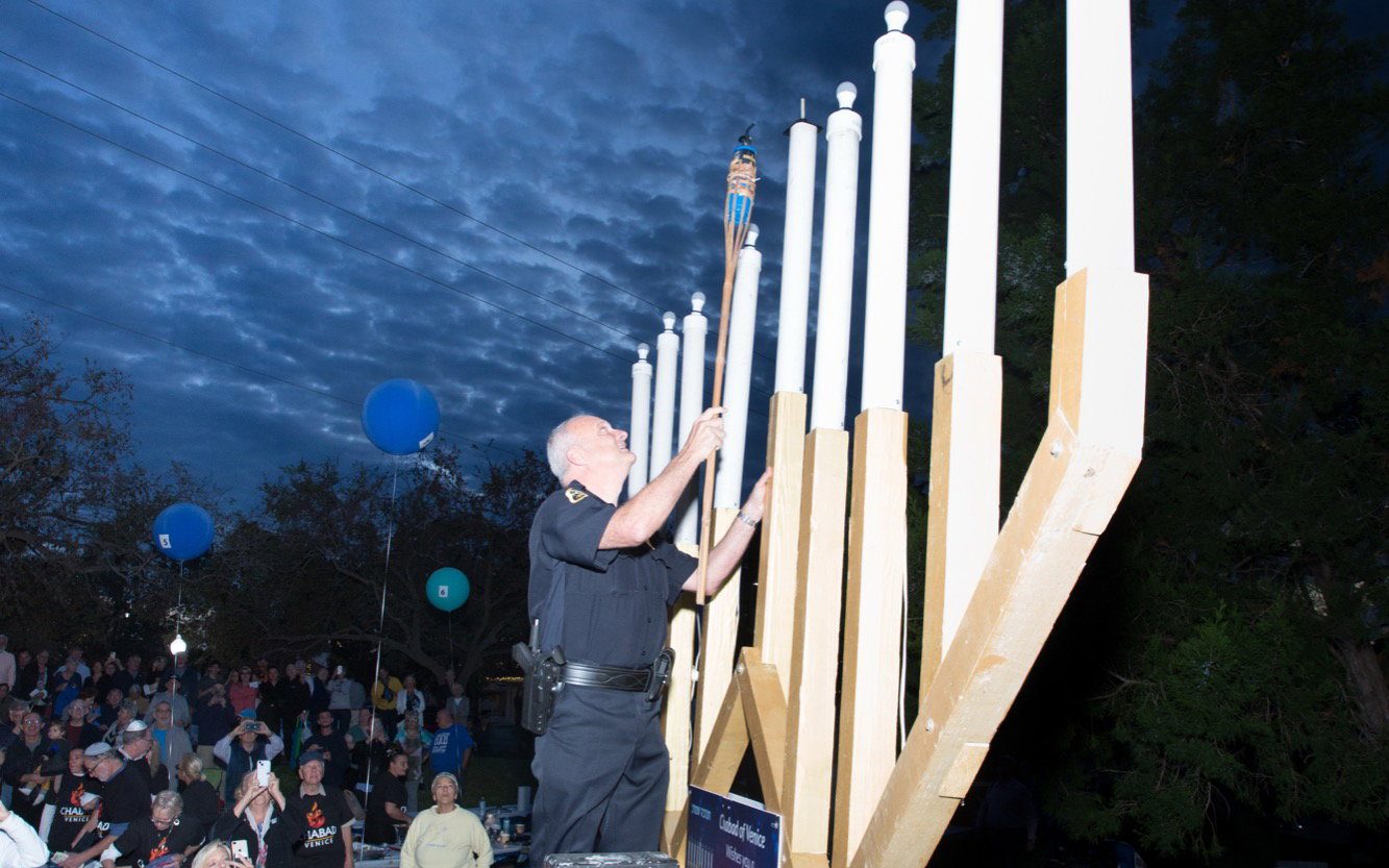 The large public menorah being lit at dusk above a seated crowd